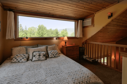 Cozy bedroom with a quilted bed, wooden ceiling, large window, and warm lighting, overlooking greenery outside.