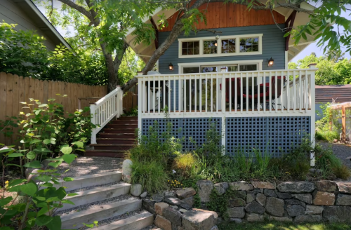 A charming blue house with a wooden deck, surrounded by greenery and stone steps leading up to it.