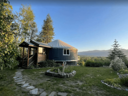 A cozy yurt surrounded by trees and a garden, with a stone path leading to a wooden entrance.