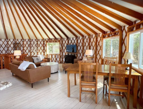 Cozy interior of a yurt with wooden beams, a seating area, dining table, and large windows letting in natural light.