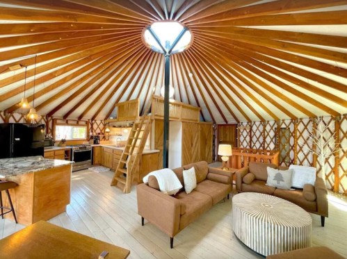 Cozy interior of a yurt with wooden beams, a kitchen area, and comfortable seating. Bright and inviting atmosphere.