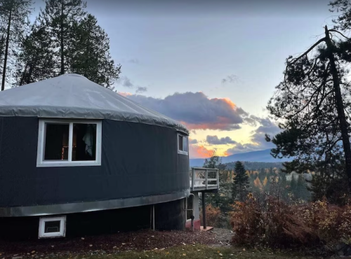 A yurt surrounded by trees, with a sunset sky and mountains in the background.