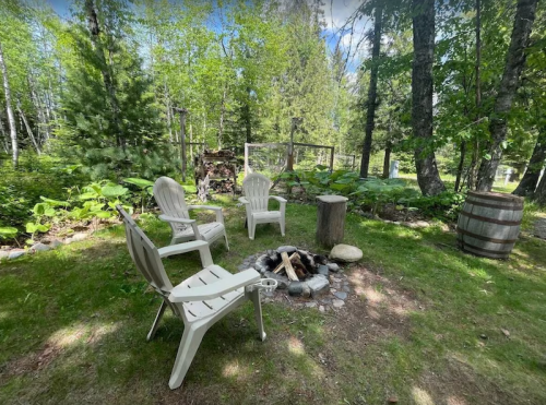 A cozy outdoor fire pit area with white chairs, a wooden stump, and lush greenery in a serene forest setting.