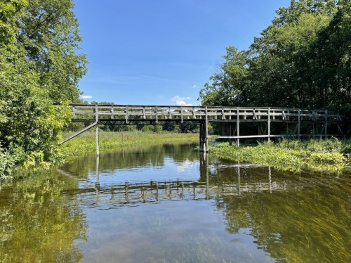 A wooden bridge spans a calm river, surrounded by lush greenery and a clear blue sky. Reflections shimmer on the water.
