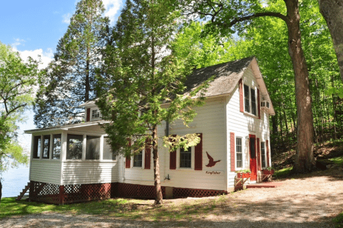 A charming white cottage with red accents, surrounded by lush green trees near a lake.