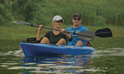 A man and woman paddle a blue kayak together on a calm waterway, surrounded by lush greenery.