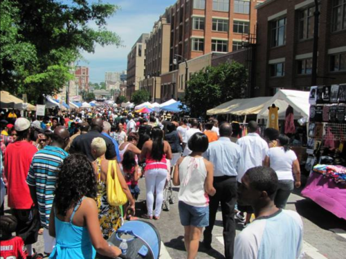 A bustling street fair with diverse crowds, vendors, and colorful tents under a clear blue sky.
