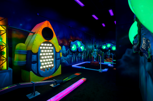 A vibrant indoor mini-golf course featuring a rocket ship and glowing decorations in a space theme.