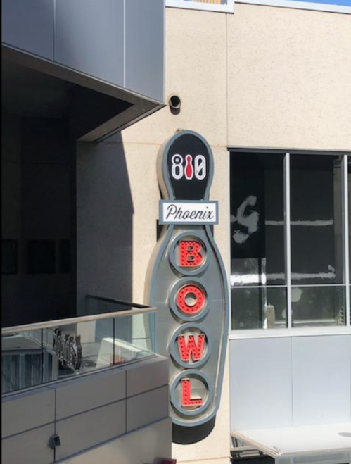 Sign for Phoenix Bowl featuring a bowling pin design and bright red letters against a modern building backdrop.