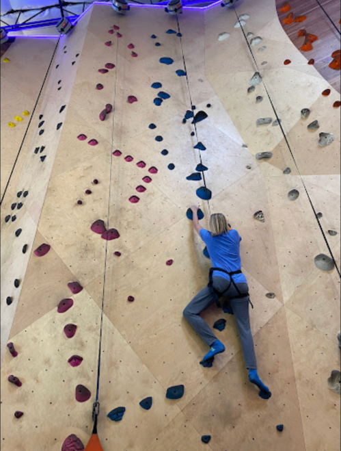 A climber ascends a colorful indoor rock wall, using various holds and wearing climbing gear.