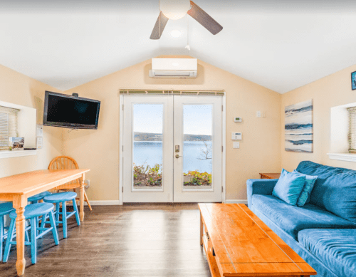 Cozy living area with a blue sofa, wooden table, and large windows overlooking a lake. Bright and inviting atmosphere.