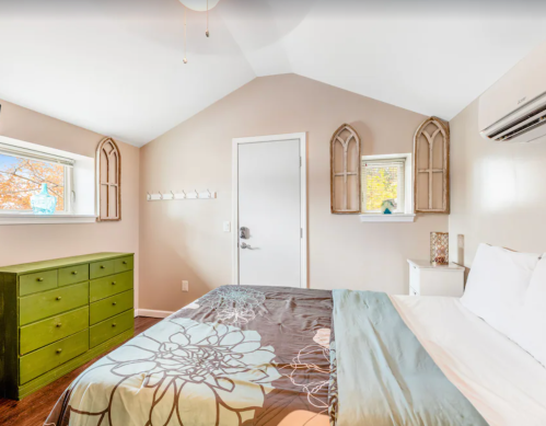 Cozy bedroom with a green dresser, white bed, and large windows, featuring light walls and decorative accents.