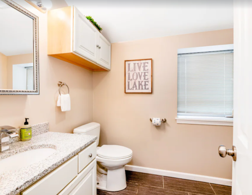 A clean bathroom featuring a sink, toilet, and a wall sign that says "LIVE LOVE LAKE." Light-colored walls and cabinets.