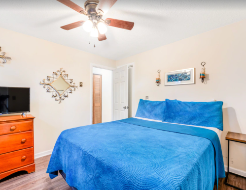 Cozy bedroom featuring a blue bedspread, wooden dresser, TV, and ceiling fan in a warm, inviting atmosphere.