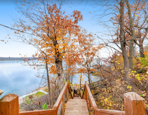 A wooden staircase leads down to a serene lake, surrounded by colorful autumn trees and clear blue skies.