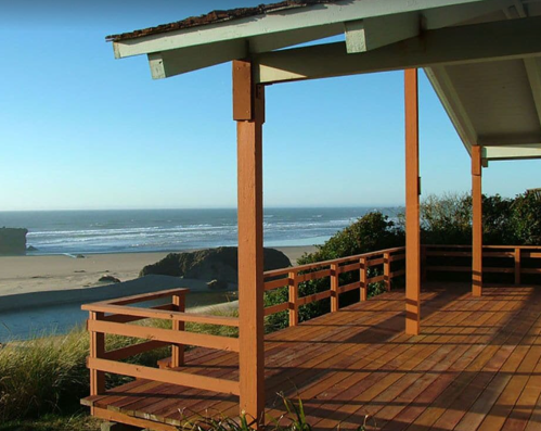A wooden deck with orange railings overlooks a sandy beach and ocean under a clear blue sky.