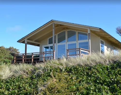 A modern house with large windows, situated on a grassy hill under a clear blue sky.