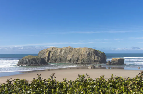 A rocky island stands in the ocean, surrounded by sandy beach and gentle waves under a clear blue sky.
