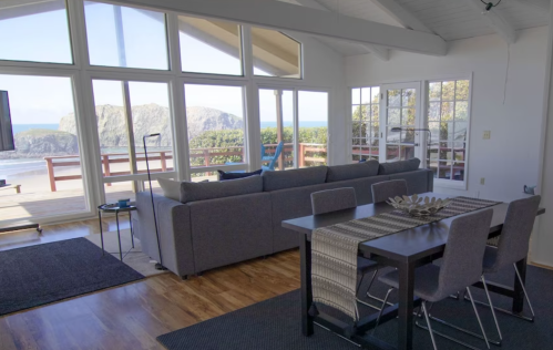 Bright, modern living space with large windows showcasing a coastal view, featuring a sofa and dining table.
