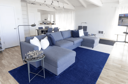 A modern living room featuring a gray sectional sofa, blue rug, and a dining area in the background with a kitchen.