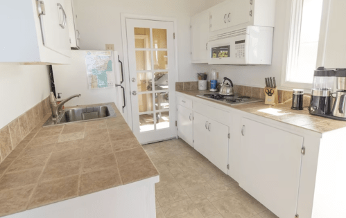 Bright kitchen with white cabinets, tiled countertops, a sink, and a door leading to an outdoor area.