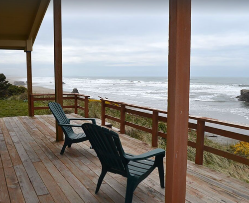 Two green chairs on a wooden deck overlook a calm beach with gentle waves and a cloudy sky.