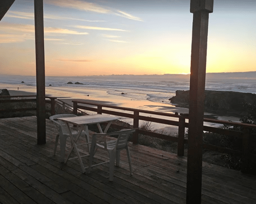 A serene beach view at sunset from a wooden deck with a table and chairs overlooking the ocean.