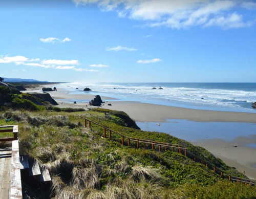 A scenic beach view with sandy shores, rocky formations, and gentle waves under a clear blue sky.