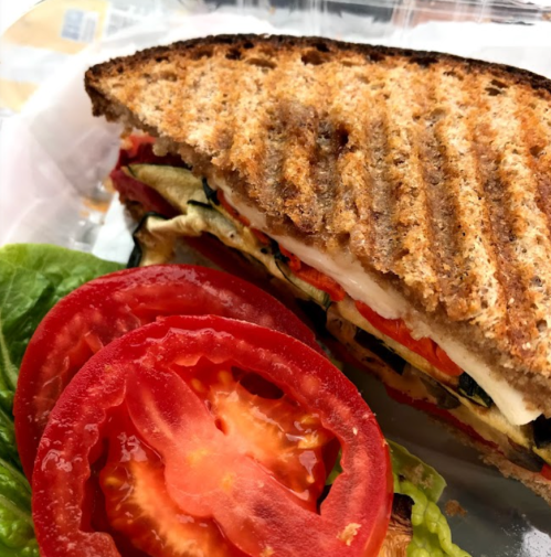 A toasted sandwich with layers of vegetables, including zucchini and tomato, served with fresh tomato slices and lettuce.