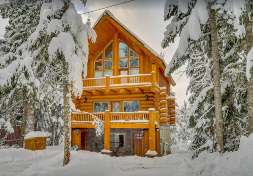 A cozy log cabin surrounded by snow-covered trees, featuring large windows and wooden balconies.