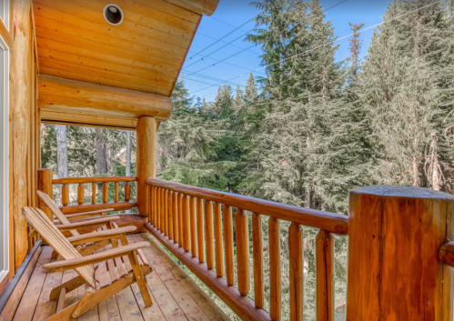 A wooden balcony with two chairs overlooks a lush forest of tall trees under a clear blue sky.