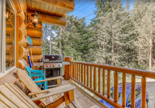 A cozy log cabin porch with wooden chairs, a grill, and a view of lush green trees.