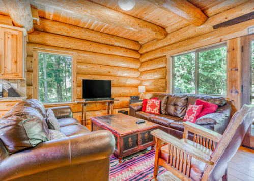 Cozy log cabin living room with leather sofas, a wooden coffee table, and large windows overlooking trees.