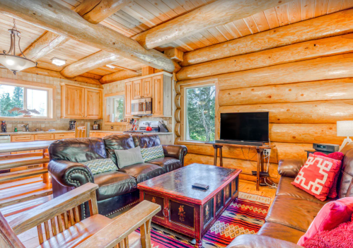 Cozy log cabin interior with wooden beams, leather sofas, a coffee table, and a TV, surrounded by large windows.