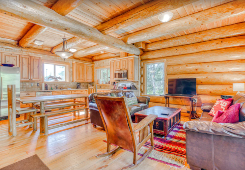 Cozy log cabin interior featuring wooden beams, a dining table, comfortable seating, and large windows.