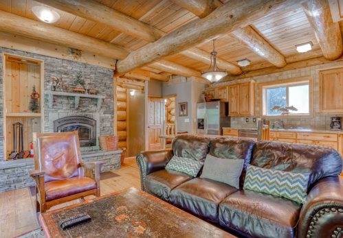Cozy rustic living room with log beams, stone fireplace, leather couch, and wooden kitchen in the background.