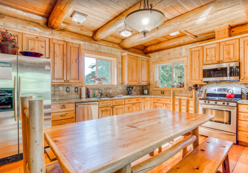 A rustic kitchen with wooden cabinets, a large table, and stainless steel appliances, featuring natural light from windows.