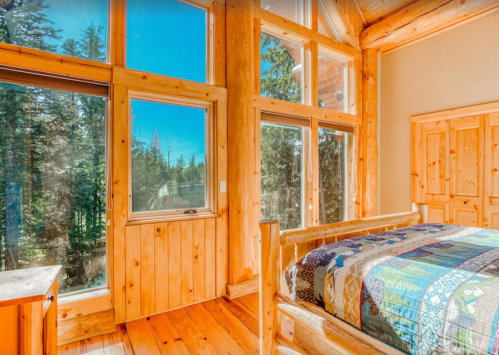 Cozy wooden bedroom with large windows showcasing a forest view and a colorful quilt on the bed.