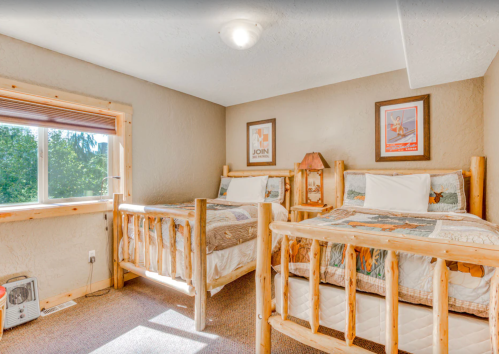 Cozy bedroom with two wooden beds, quilted bedding, and a window overlooking greenery. Warm, inviting decor.