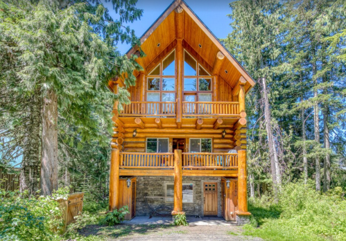 A large log cabin with a peaked roof, balconies, and surrounded by tall trees in a lush green setting.