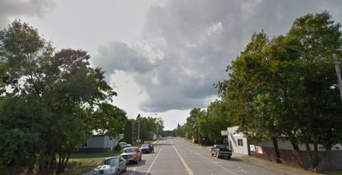A quiet street lined with trees, featuring parked cars and a cloudy sky overhead.
