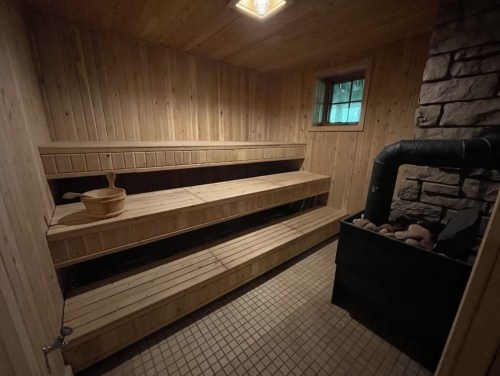 A wooden sauna interior with benches, a stone wall, and a stove with rocks, illuminated by soft lighting.