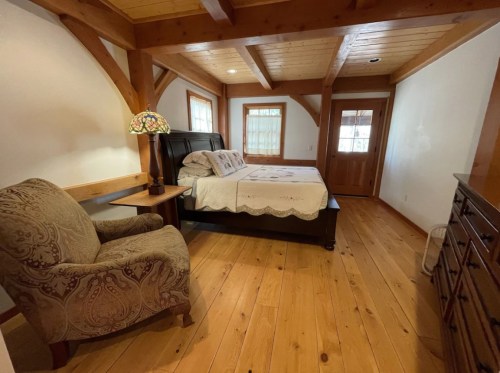 Cozy bedroom with wooden beams, a bed with a quilt, a patterned chair, and a dresser in a warm, inviting atmosphere.