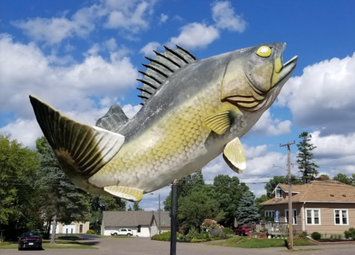A large, colorful fish sculpture mounted on a pole against a blue sky with clouds, near a residential area.