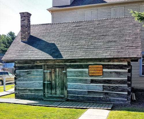 A rustic log cabin with a chimney and a wooden sign, set against a backdrop of a larger building.
