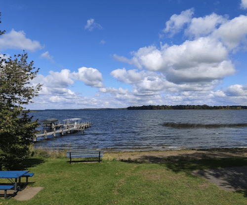 A serene lakeside view with a dock, grassy area, and fluffy clouds in a blue sky.