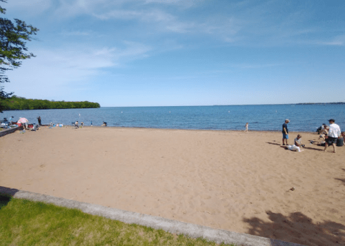 A sandy beach by a calm lake, with people enjoying the sun and water on a clear day.