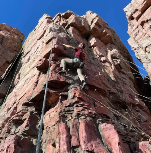 A climber ascends a rocky cliff under a clear blue sky, secured by climbing ropes.