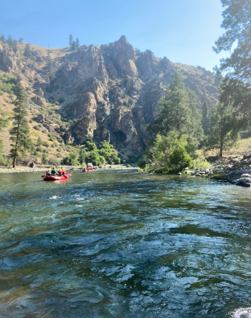 A scenic river flows through rocky terrain, with trees lining the banks and a group rafting in the distance.
