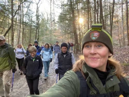 A group of hikers walking on a forest trail, with a woman in the foreground smiling at the camera.